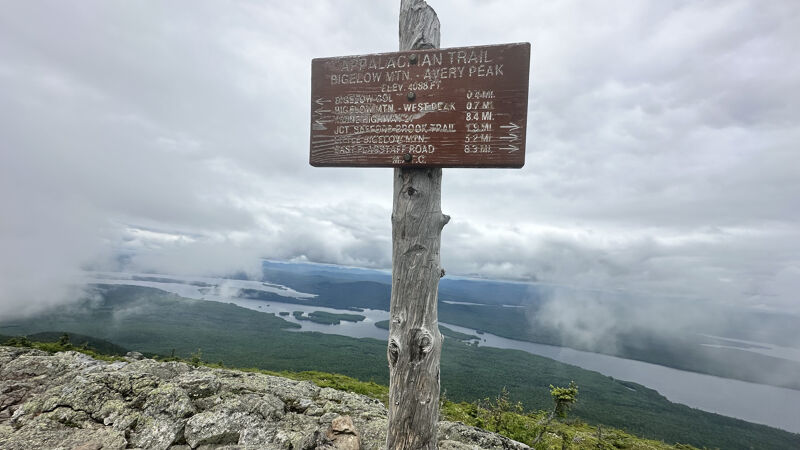 The image shows a wooden signpost on a mountain, with a cloudy sky in the background. The sign indicates the directions and distances to various trails, including the Appalachian Trail. The landscape features a view of a lake and green vegetation. The overall scene suggests a hiking trail in a natural environment.